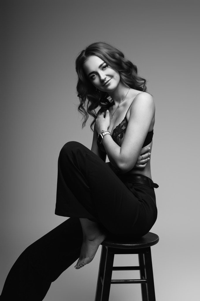 Kõhn naine istub taburetil, poseerib mustvalge fotosessiooni jaoks, elegantne stiil ja pehme valgustus.