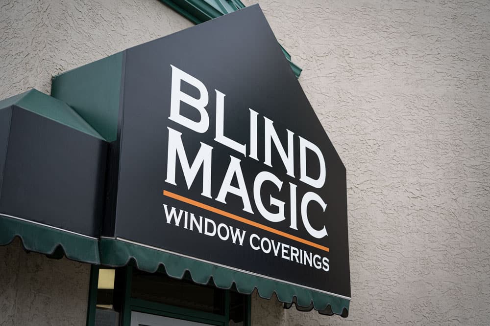 Bright black and white sign displaying "Blind Magic Window Coverings" on a storefront, promoting expert window covering solutions.