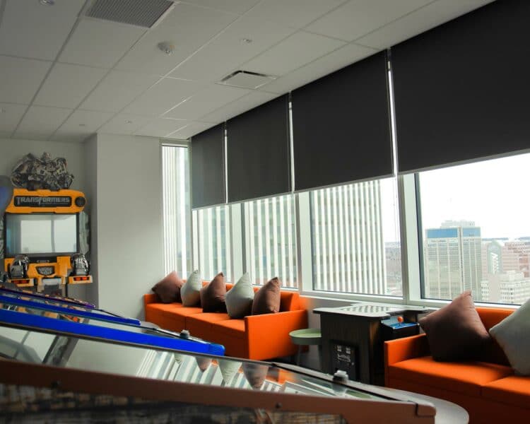 Bright modern lounge with blackout blinds, orange sofas, and gaming arcade in a contemporary office setting.