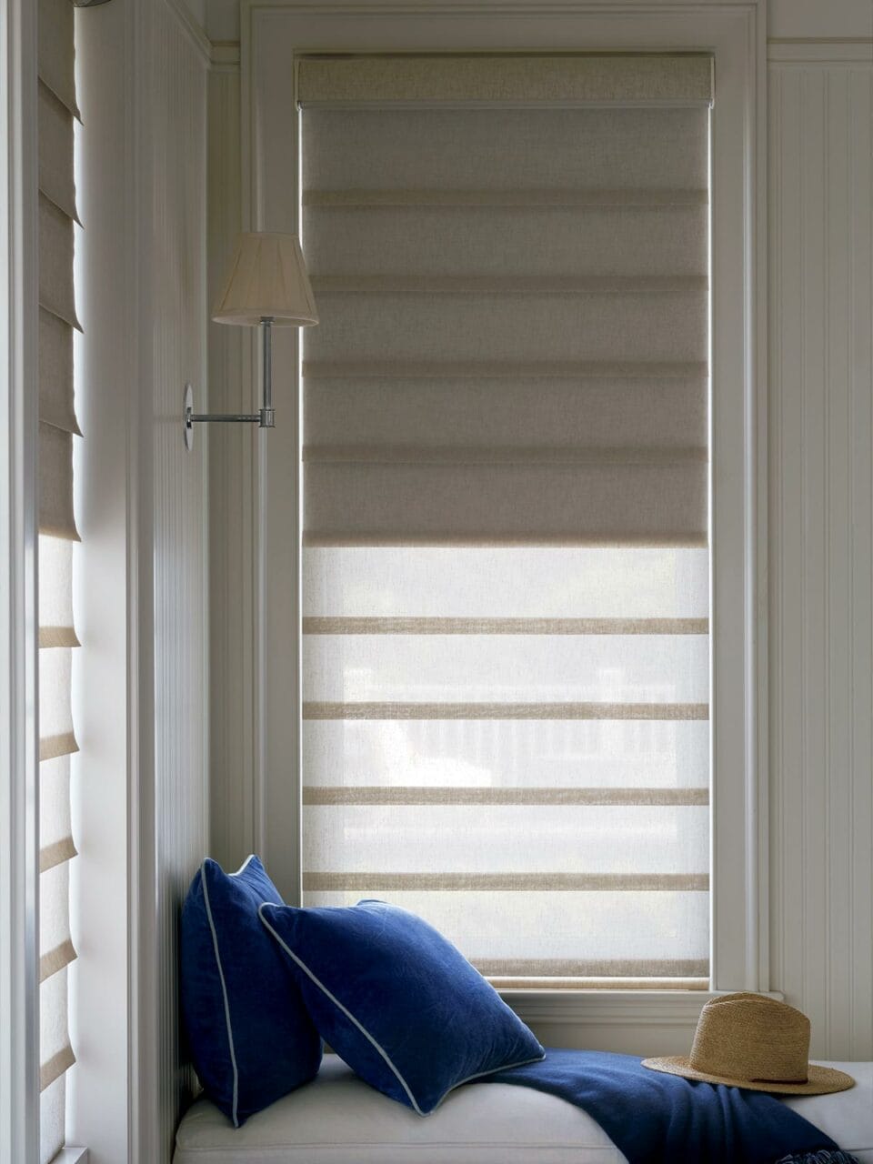 Soft beige window blinds with a cozy blue cushion and straw hat by the window, creating a relaxing reading nook.