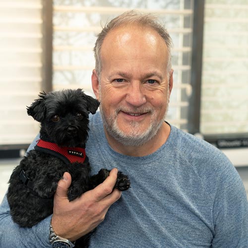 Man smiling with a small black dog in his hand.