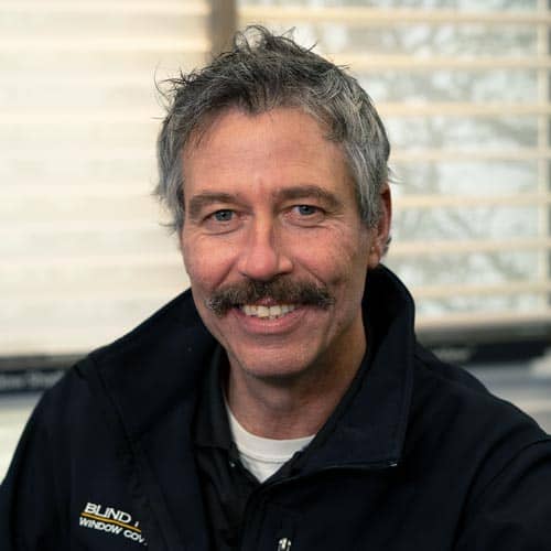 Experienced man in a black jacket with "Blind Window" logo, smiling in an office setting with window blinds.