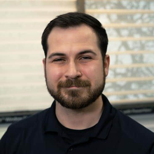 Man with a beard and short dark hair smiling indoors, casual black shirt, bright natural light background.