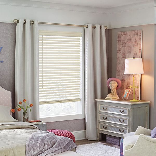 Bright, cozy bedroom featuring large window with white blinds and curtains, elegant white dresser with decorative bust, soft lighting, and tasteful wall art.