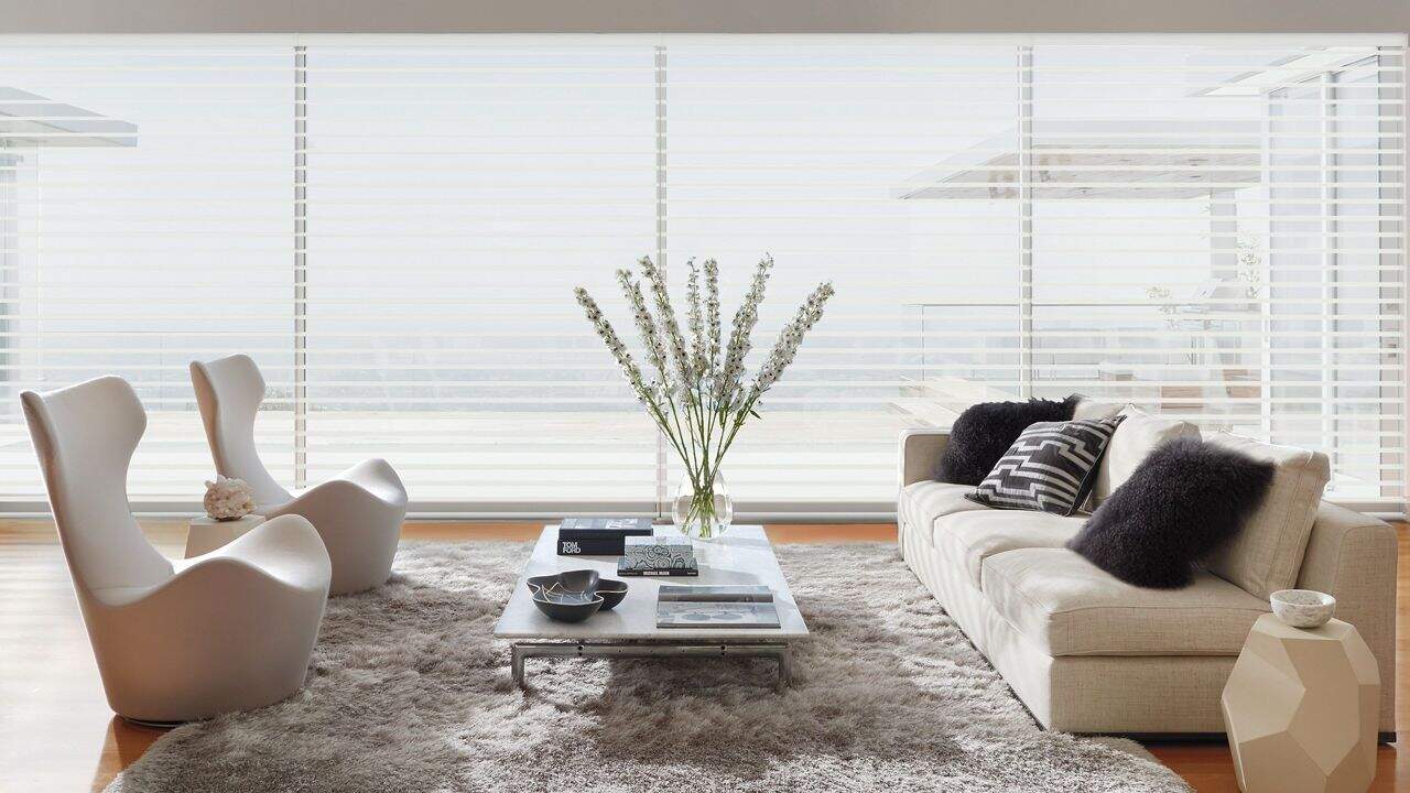 Bright living room with beige sofa, white chairs, and glass coffee table in front of floor-to-ceiling window blinds.