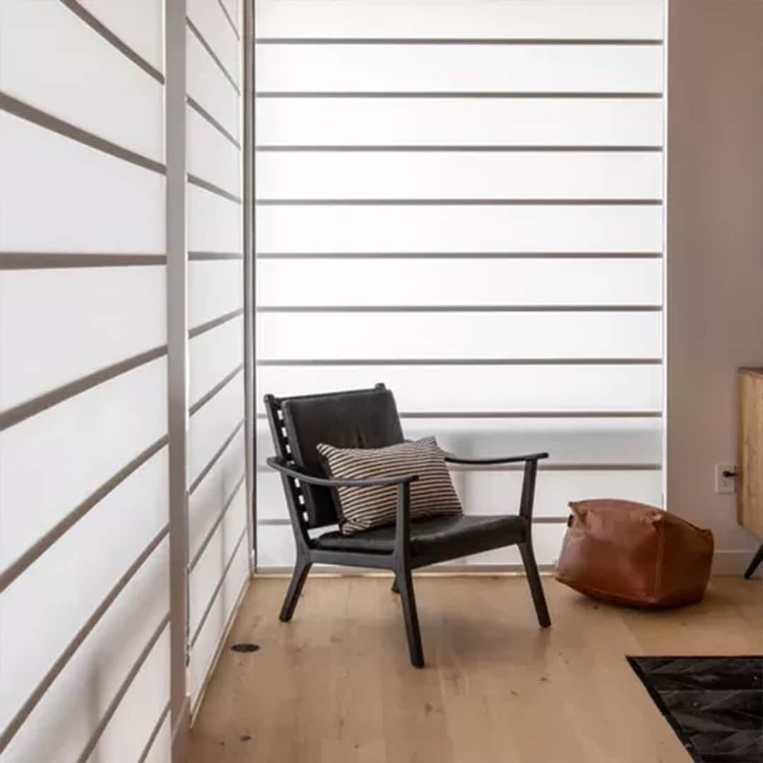 Stylish black lounge chair with striped pillow placed in a well-lit modern room featuring large white window blinds.