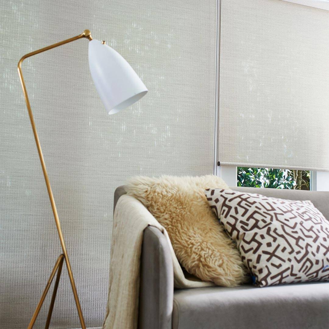 A comfortable living room corner featuring a beige sofa with decorative pillows, a cozy blanket, and a stylish floor lamp against a window with natural light and green views.