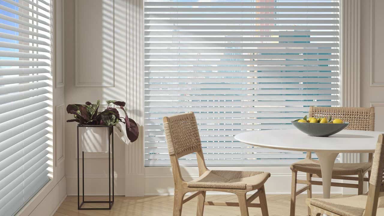 Bright dining area with large windows and horizontal blinds, natural light, modern decor, wooden chairs, round table, bowl of lemons, minimalistic interior, cozy yet elegant vibe, indoor sunlight.