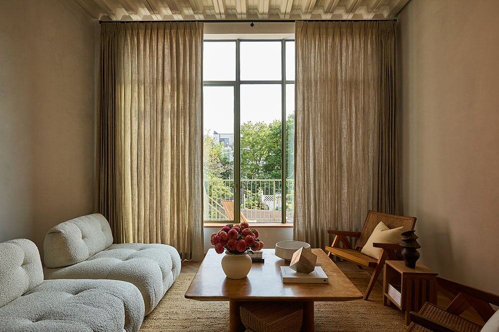 Bright living room with large window and natural lighting, plush white sofa, wooden coffee table with decorative items, rattan chair with cushion, beige curtains, and green outdoor view.