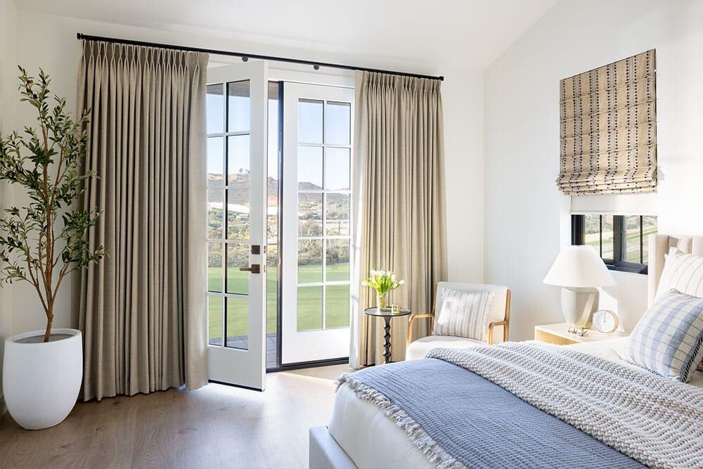 Bright bedroom featuring French doors, large windows, and elegant curtains enhancing natural light and outdoor views. Modern decor with a bed, armchair, side table, and potted plant creates a relaxing retreat.