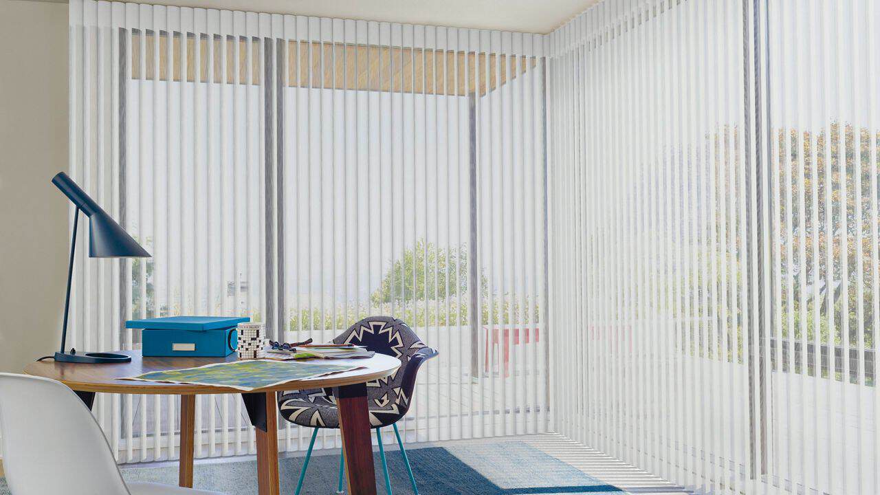 Vertical blinds in a modern bright room with large windows, providing privacy and light control. Minimalist decor with a small table, chair, and outdoor view.