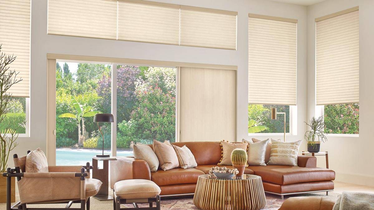 Spacious living room featuring large windows with beige blinds, a tan leather sofa, contemporary decor, and views of greenery outside. Bright, airy, and stylish for relaxing or entertaining.