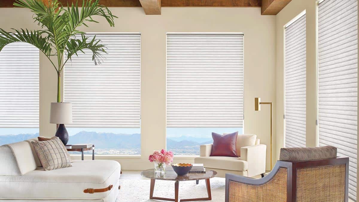 Modern sunlit living room with large windows and white blinds, stylish furniture, potted plant, mountain view, cozy decor, neutral colors, natural light, comfortable seating, elegant interior design, relaxing ambiance.