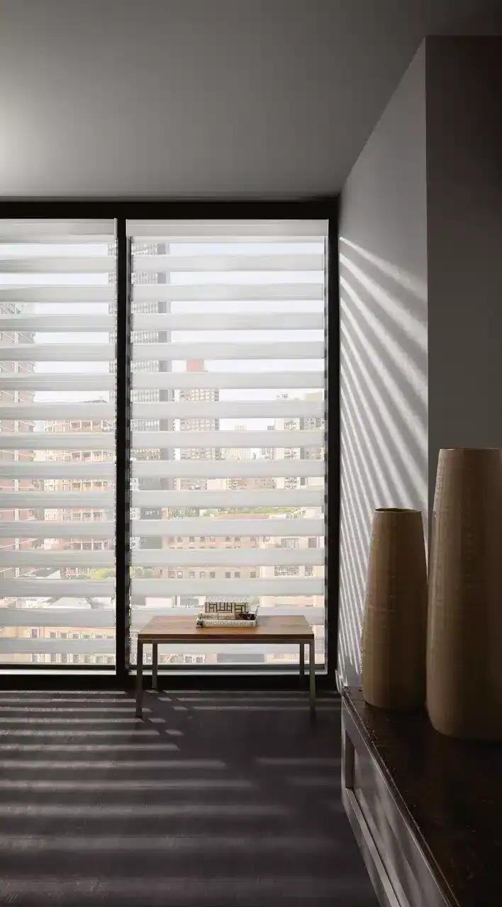 Modern apartment window with horizontal blinds casting light and shadows, city skyline visible outside.