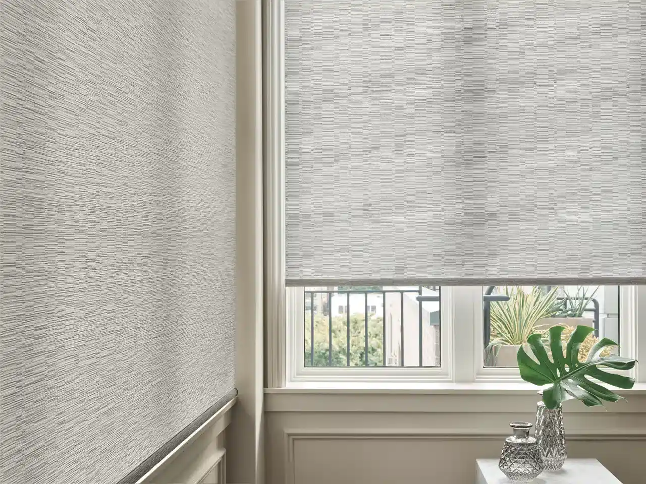 ALT text: Interior window scene with textured roller blinds, a small table with decorative vases and a Monstera leaf, framed by a classic white window and wall trim.