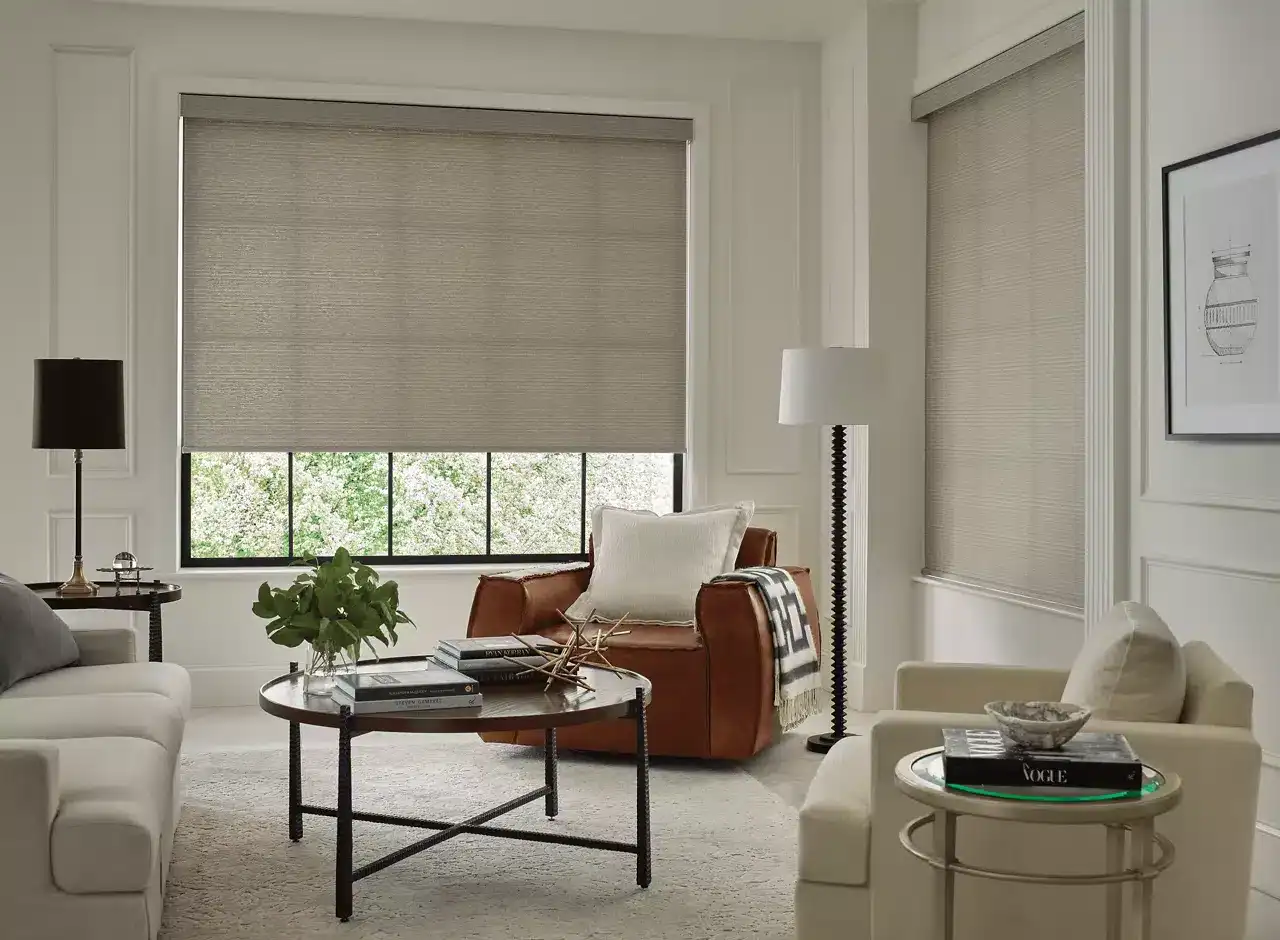Bright and inviting living room with large windows, beige blinds, and elegant furniture including white sofas, a leather armchair, and stylish side tables.