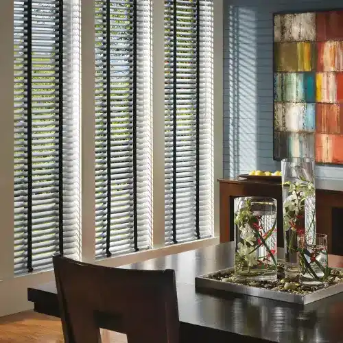 Bright sunlight streams through window blinds in a stylish dining area with artistic wall decor and a floral centerpiece, creating a welcoming and elegant atmosphere.