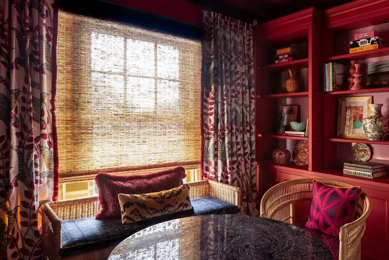 Bright window with bamboo blinds, floral curtains, and colorful cushions create a warm, inviting space for reading or relaxing.
