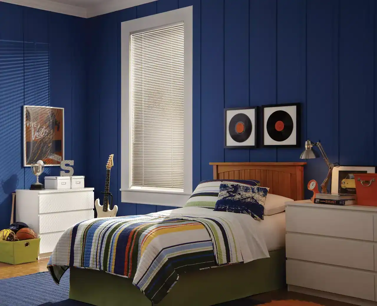 Bright blue bedroom wall with vinyl record art, a white dresser, skateboard, and sports gear, creating a vibrant and lively ambiance. Natural light filters through blinds, illuminating the colorful bedrooms' decor perfect for a creative personality.