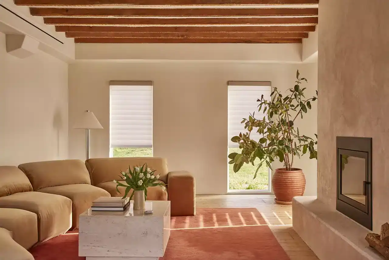 Bright living room featuring a modern fireplace, large windows with white blinds, a beige sofa set, indoor plants, and wooden ceiling beams for a warm, inviting atmosphere.