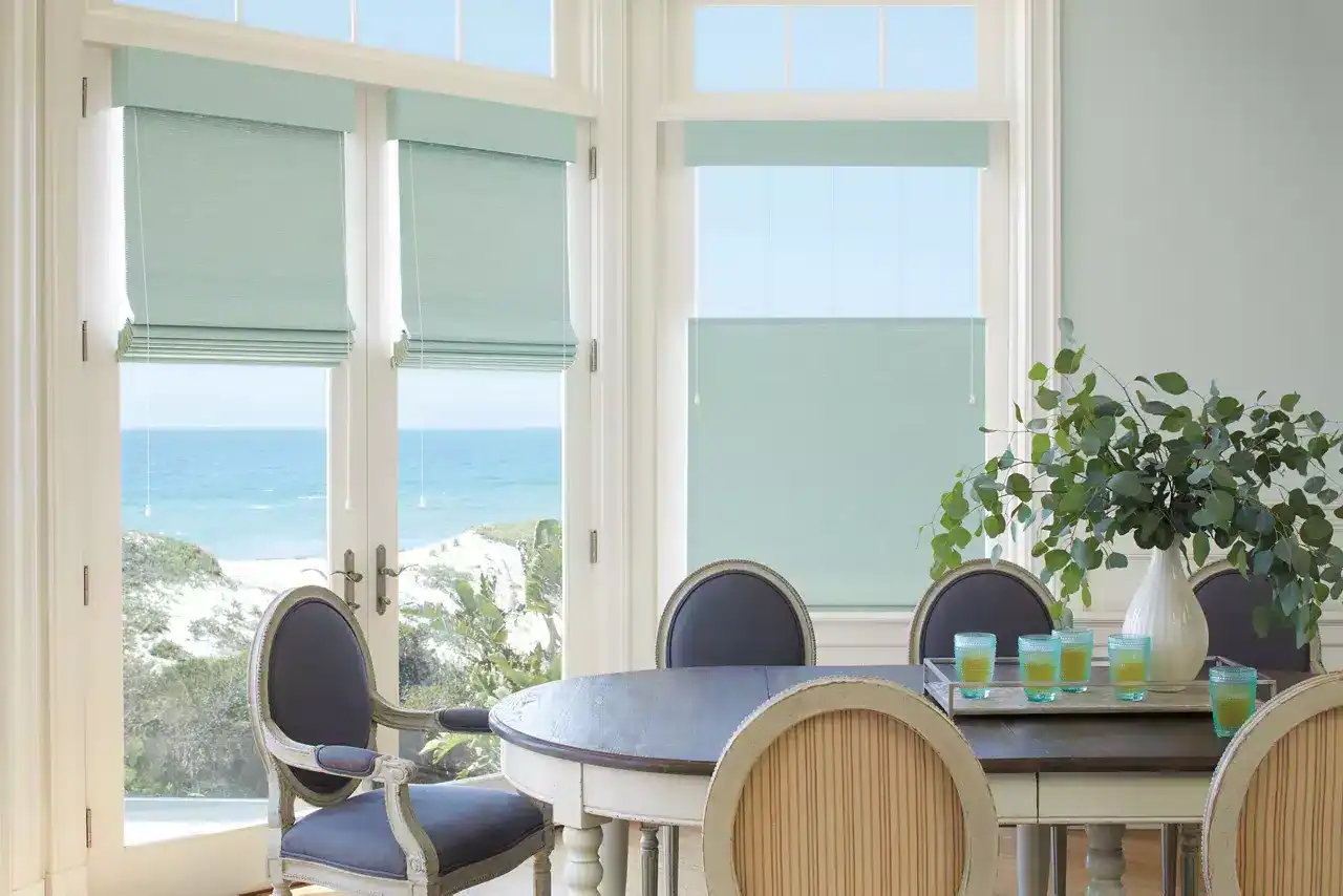 Bright seaside dining area featuring large windows overlooking the ocean with blue shades and greenery, elegant furniture, and table settings for a relaxing coastal meal.