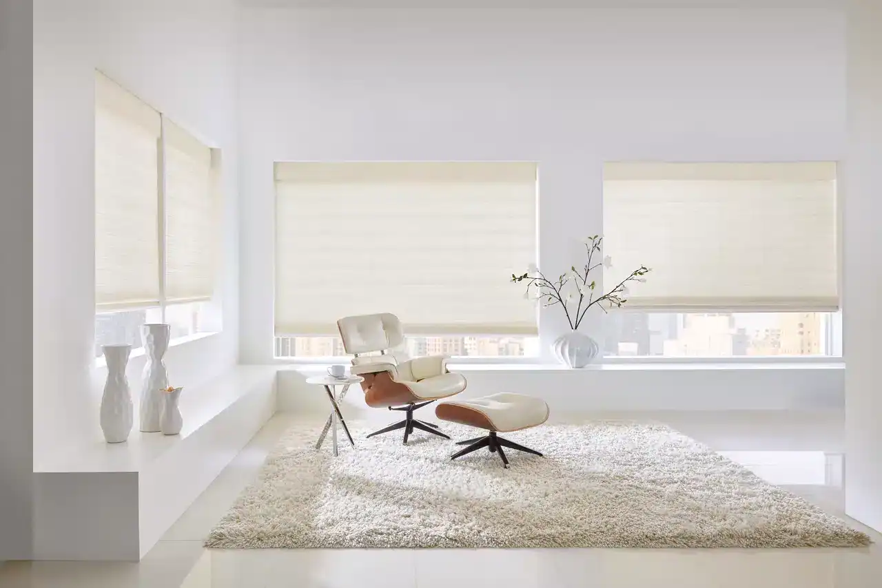 Bright and airy interior with large windows featuring cream roller blinds, modern beige lounge chair with matching ottoman, plush white rug, and elegant white vases with minimal decor.