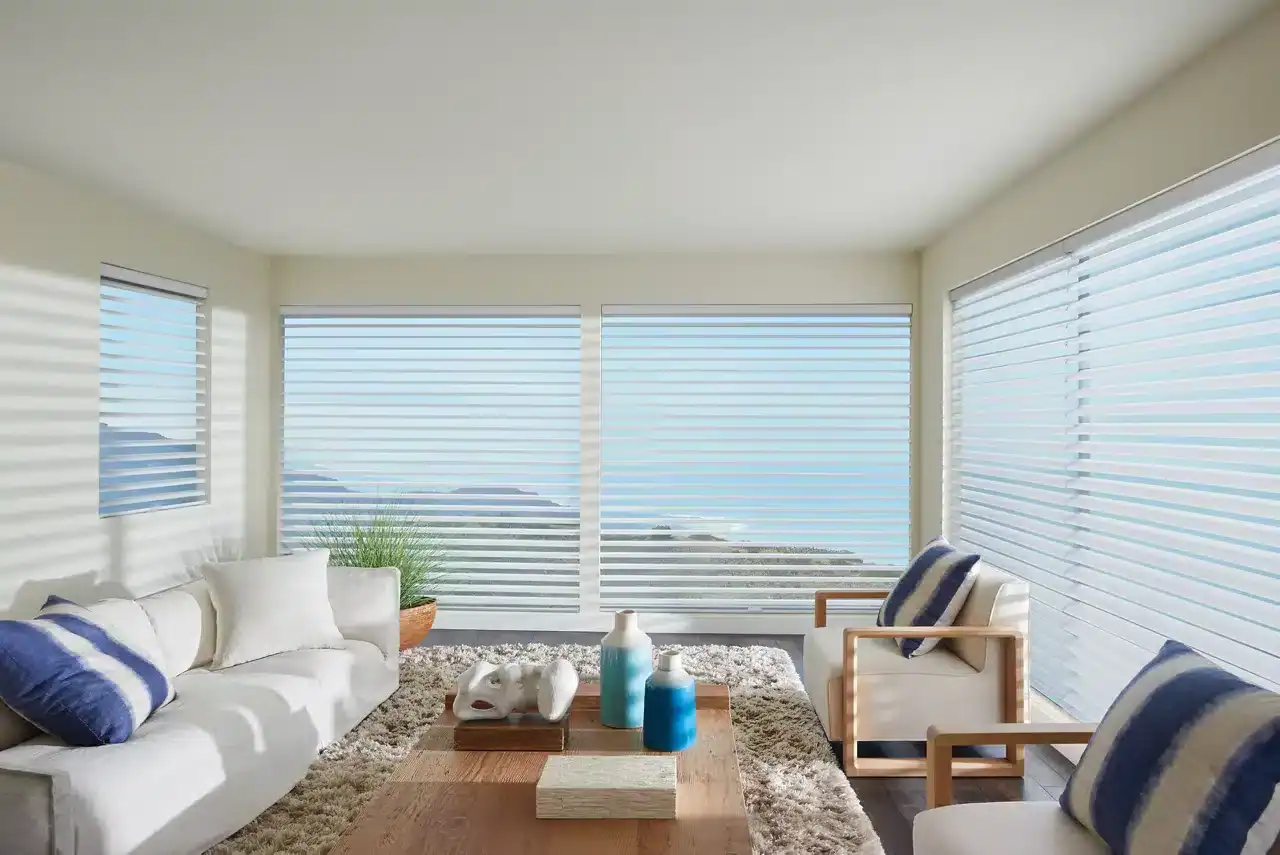 Bright living room with panoramic ocean view, white furniture, and horizontal blinds.
