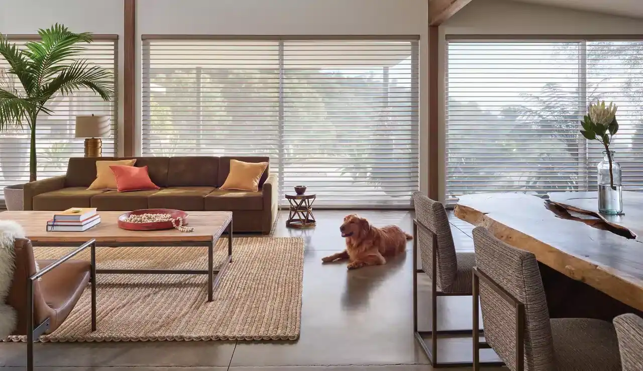 Bright living room with large windows, stylish furniture, a dog lying on a hardwood floor, lush plants, and decorative accents, creating a warm and inviting atmosphere.