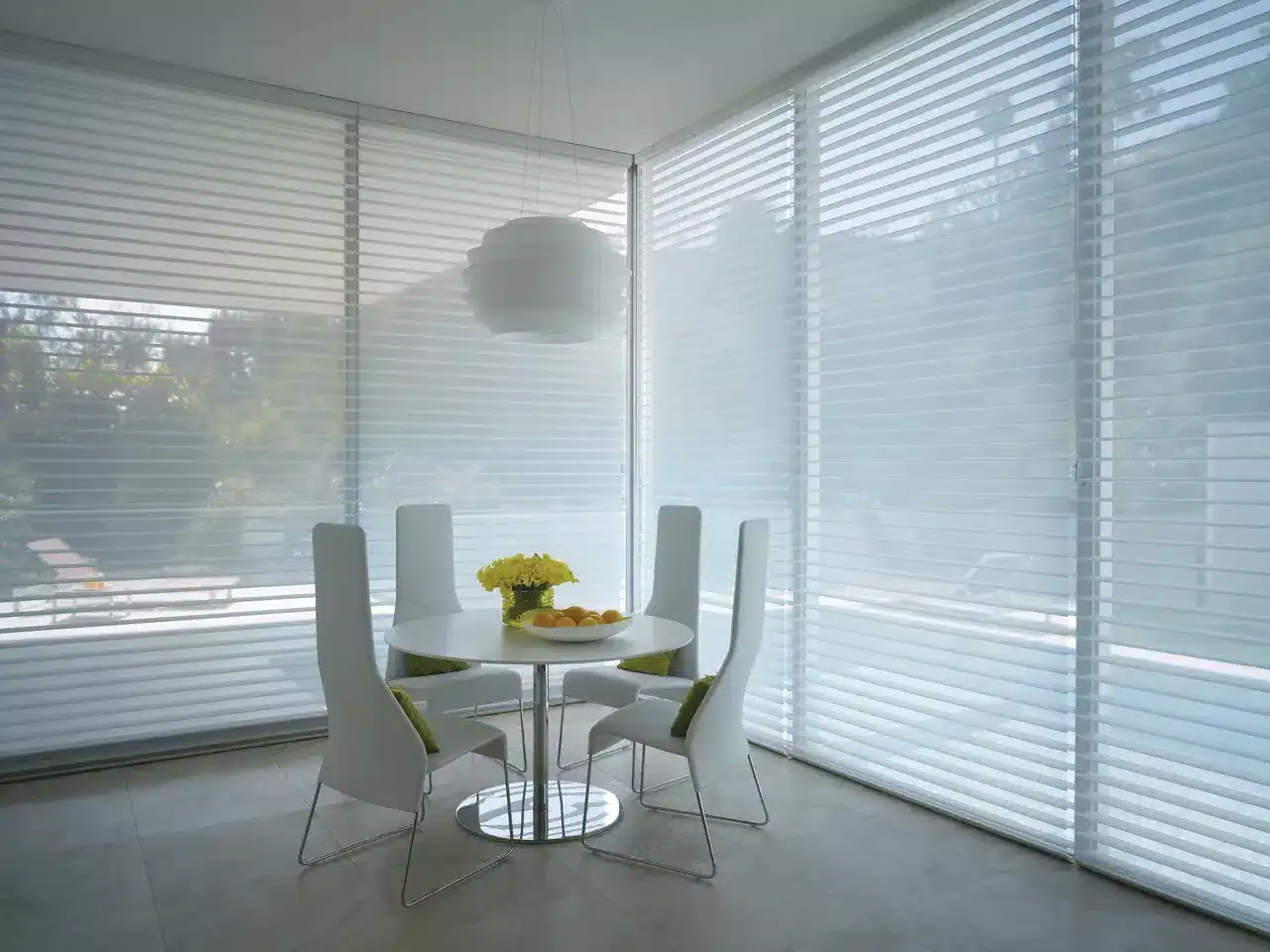 Bright, airy breakfast nook with large windows, white chairs, and a round table decorated with flowers and fruit.