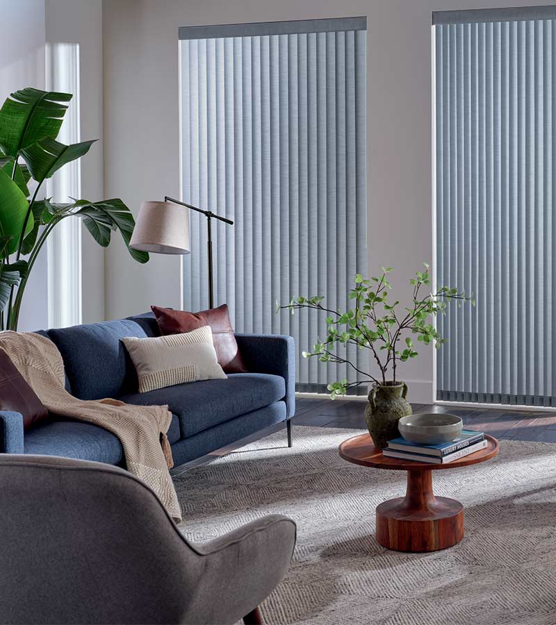 Bright living room with large windows, vertical blinds, lush green plants, modern sofa, and minimalist wooden coffee table, creating a stylish and relaxing space.