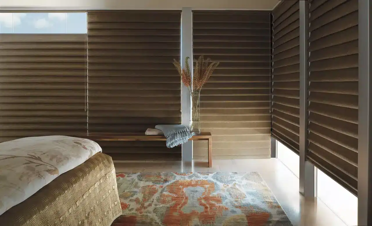 Close-up of a bedroom with brown horizontal window blinds, a bed with floral bedding, a wooden bench with a vase of dried flowers, and a colorful rug creating a warm, modern ambiance.