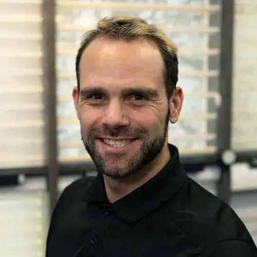 Professional portrait of Sandi Switzer, smiling man with short brown hair and beard, wearing a black shirt, in an office setting with blurred background.