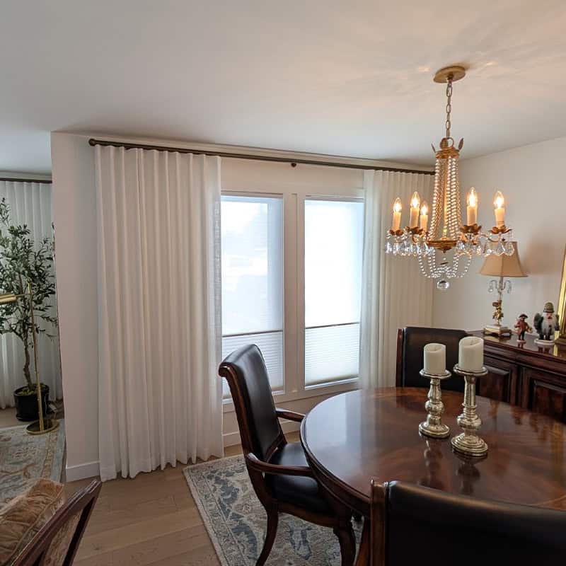 Elegant living room with large windows, white curtains, and a classic chandelier. The space features a wooden dining table, candles, and decorative elements creating a cozy, sophisticated atmosphere.