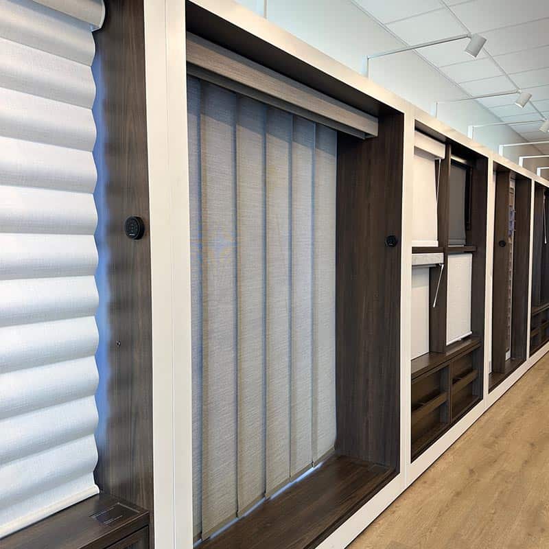 A modern showroom featuring different window treatment options, including roller blinds, vertical blinds, and wooden shutters, arranged neatly for customer selection.