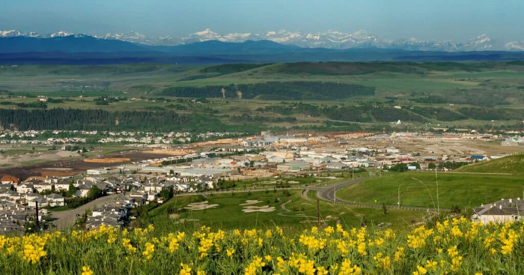 Vast landscape view of a green valley, residential area, and distant snow-capped mountains under clear blue sky.