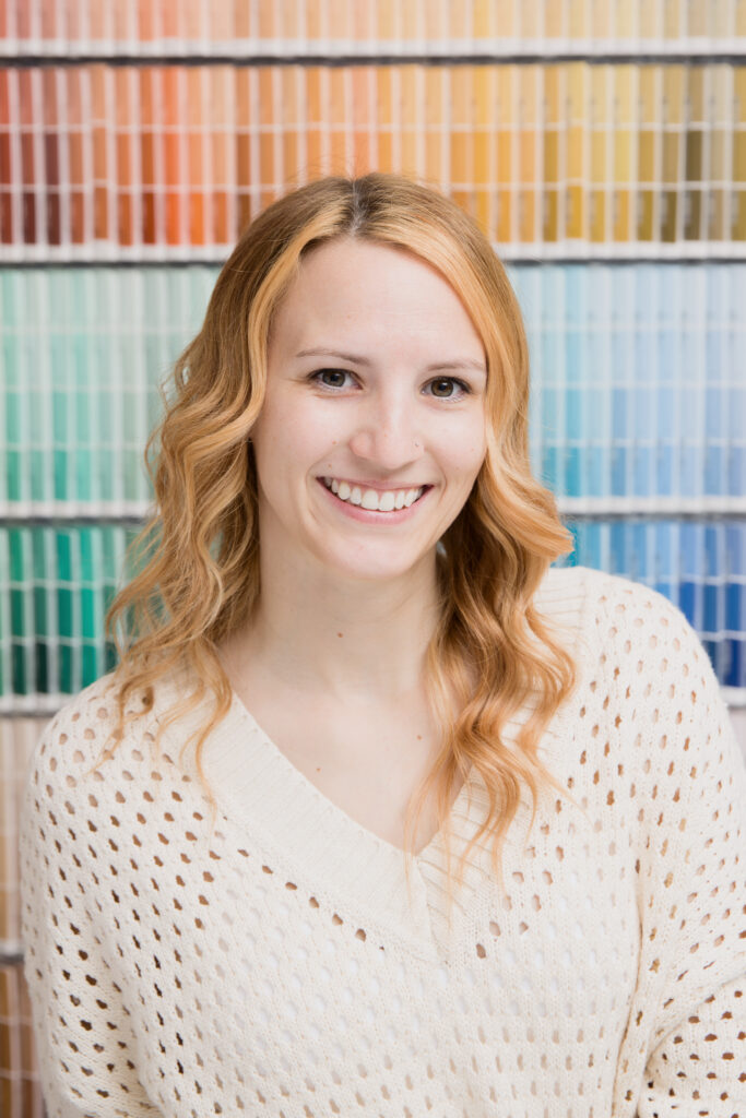 A cheerful woman with red hair and a warm smile, standing in front of a colorful paint sample display. relaxed and friendly, showcasing vibrant personality and style.