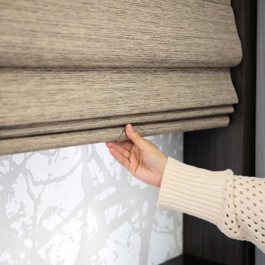 Close-up of hand adjusting beige fabric window blind with textured details, mounted on dark wood frame, featuring modern, stylish window treatment for interior decor.