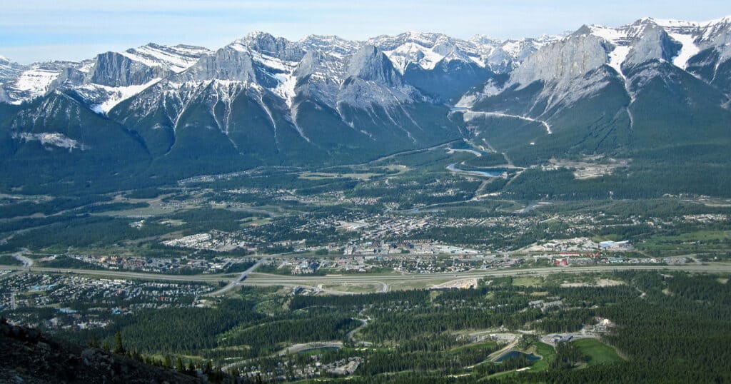 Scenic view of a mountain valley with snow-covered peaks, dense forests, and a winding road, showcasing natural landscapes and outdoor adventure opportunities in the mountainous region.