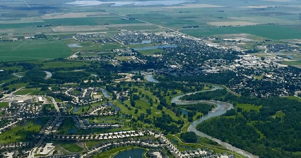 Aerial view of a suburban community featuring lush parks, winding rivers, residential neighborhoods, and agricultural fields in the background.