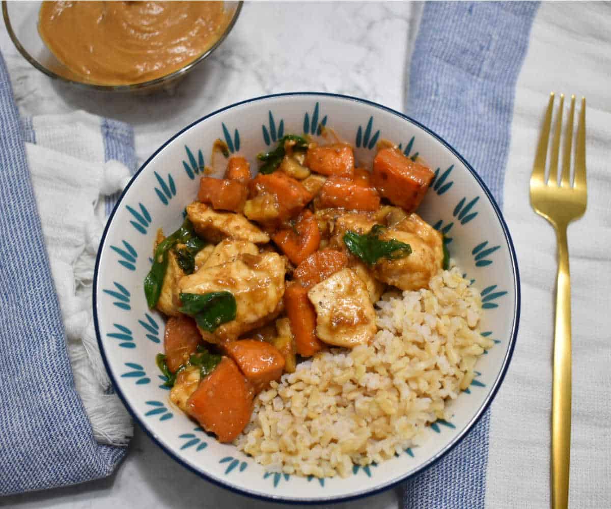 Bowl of chicken stir fry with peanut sauce, sweet potatoes, spinach, and brown rice, served on a white and blue plate with a gold fork and a side of peanut sauce.