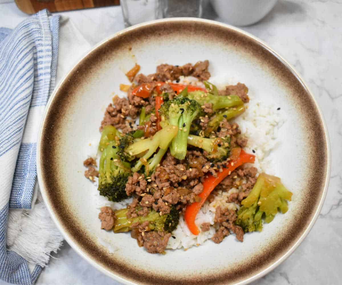 A bowl of ground beef stir fry with broccoli and red bell peppers served over white rice, topped with sesame seeds.