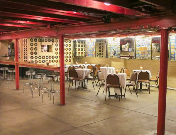 A retro-style basement lounge with tables and chairs covered in white tablecloths, red support beams, bar stools, and walls decorated with records and framed photos or posters.