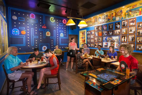 A group of people dine and chat in a retro-themed restaurant decorated with a large Pac-Man mural and framed photos on the walls. An arcade game table and a waitress are visible in the lively, colorful scene.