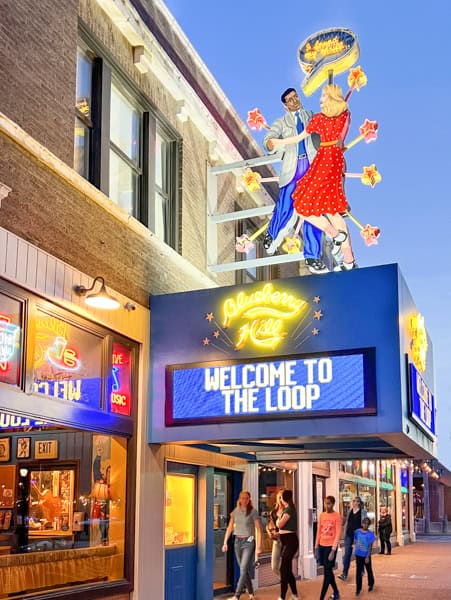People walk under a bright Blueberry Hill sign featuring a dancing couple and stars above a marquee that reads WELCOME TO THE LOOP, on a lively city street at dusk.
