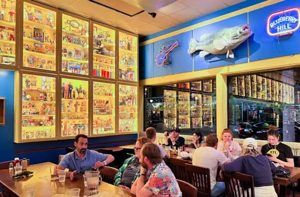A group of people sits at tables in a brightly lit restaurant with display cases full of colorful figurines and collectibles on the walls. A large fish and neon sign reading Blueberry Hill are mounted above.