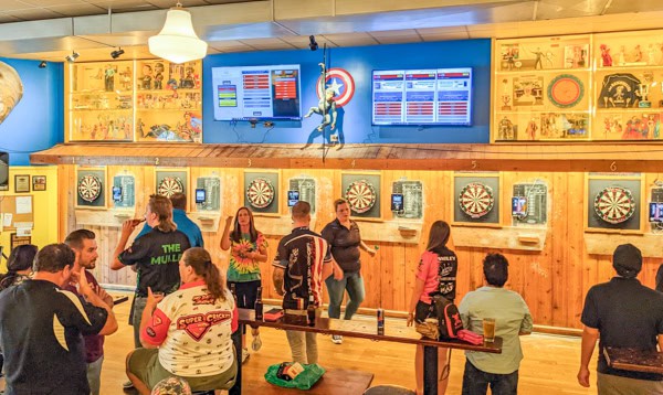 A lively bar scene with people playing darts at multiple dartboards on a wooden wall. Some people are watching, others are throwing darts. Colorful scoreboards and decorative art are visible on the walls.