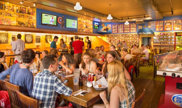 A lively bar and restaurant with people eating, drinking, and socializing. Several groups sit at wooden tables, while others stand by dartboards. The walls are decorated with pictures, memorabilia, and TVs showing sports.