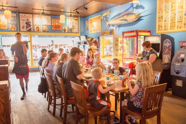 A group of people, including children and adults, sit around a table eating in a colorful, casual restaurant decorated with posters, a large mounted fish, and arcade machines. A waiter carries food toward the group.