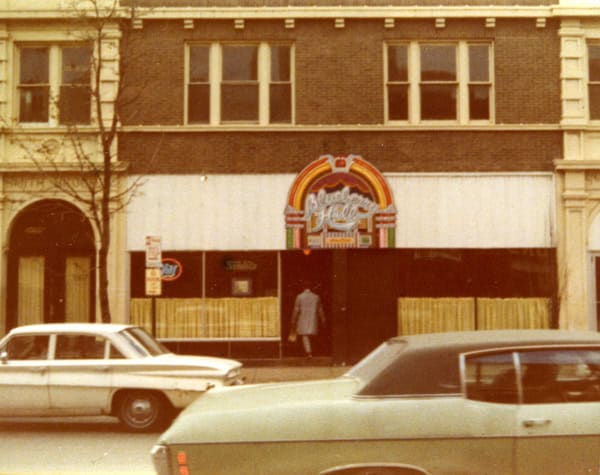 A vintage photo of a street with two classic cars parked in front of a brick building. The building has large windows above and a colorful “Peaches” sign on the ground floor. A person is walking toward the entrance.