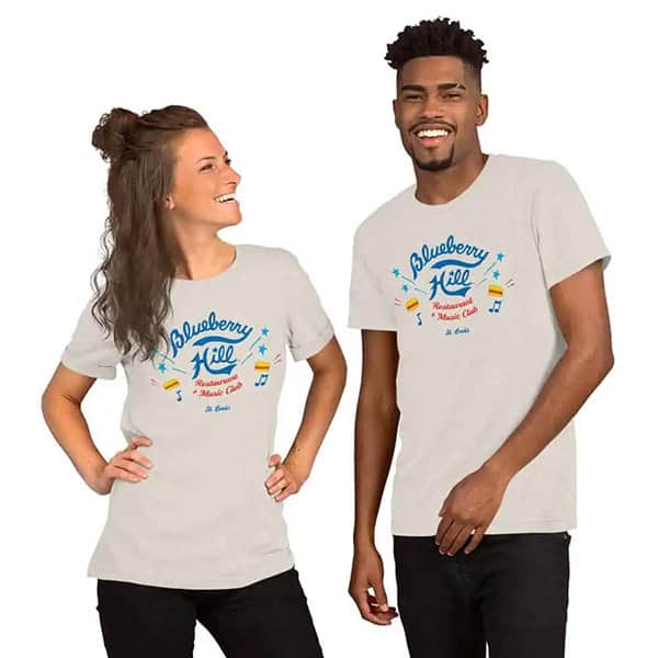 Two people wearing matching light gray Blueberry Hill graphic t-shirts stand smiling, with one holding their hip and the other with a hand in their pocket, against a plain white background.